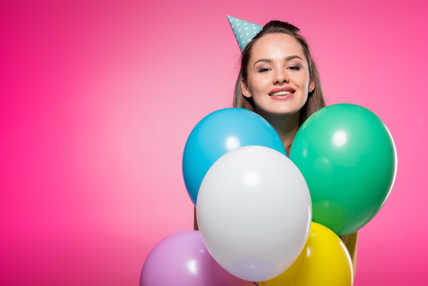 attractive woman with party hat looking out from balloons isolated on pink - Fotografie, Obrázek