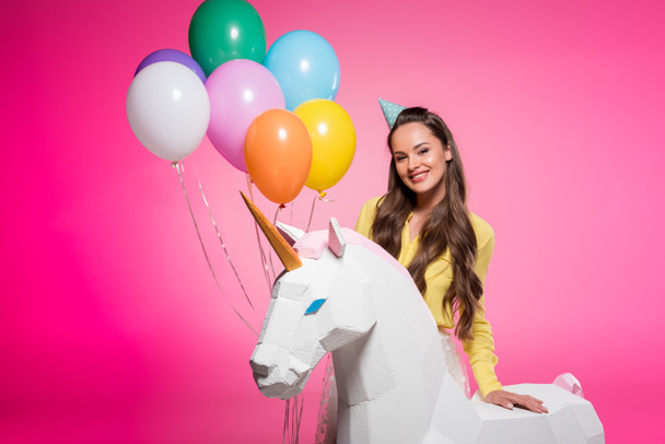 atractiva mujer con sombrero de fiesta, globos y unicornio juguete aislado en rosa
 - Foto, Imagen