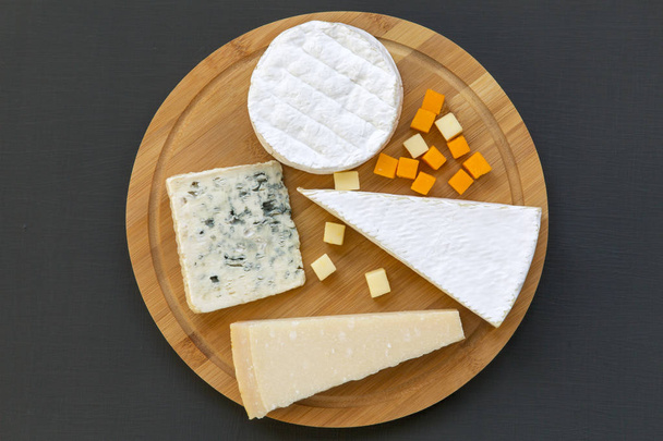 Tablero de madera con varios tipos de queso sobre fondo oscuro, vista superior. Acostado. Desde arriba
. - Foto, Imagen