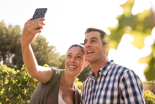 A man and woman taking a selfie in the vineyards smiling broadely at the phone camera - Foto, afbeelding