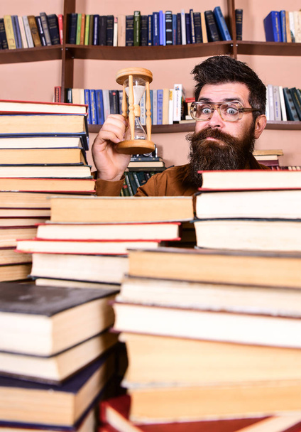 Man on surprised face holds hourglass while studying, bookshelves on background. Time flow concept. Teacher or student with beard studying in library. Man, scientist in glasses looks at hourglass - Photo, image