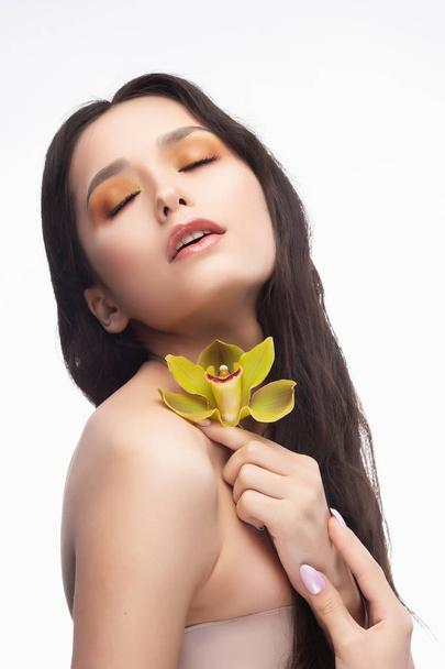 A beautiful brunette nude shoulders girl sensually holds a flower of an orchid in her hands. Nude make-up. Healthy smooth skin. Isolated on white background. Copy space. Commercial and advertising. - Фото, зображення