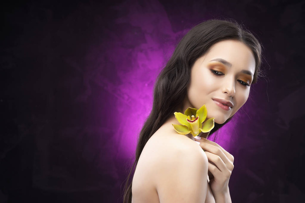 A beautiful brunette nude shoulders girl sensually holds a flower of an orchid in her hands. Nude make-up. Healthy smooth skin. Purple-black background. Copy space. Commercial and advertising. - Fotoğraf, Görsel