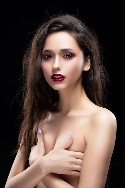 Portrait of a beautiful glamorous brunette girl sensually covers her naked breast with her hands. Shimmering makeup, red lips. Healthy smooth skin. Isolated on black. Copy space. - Fotografie, Obrázek