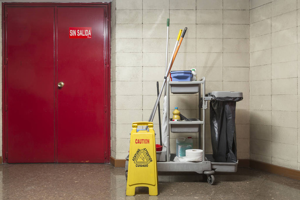 Professioneller Reinigungswagen, ausgestattet mit Produkten und Werkzeugen, sowie ein großer Müllsack, der neben einem tragbaren Warnschild für nasse Böden platziert ist, auf der anderen Seite eine rote Tür mit einem "No exit" -Schild - Foto, Bild