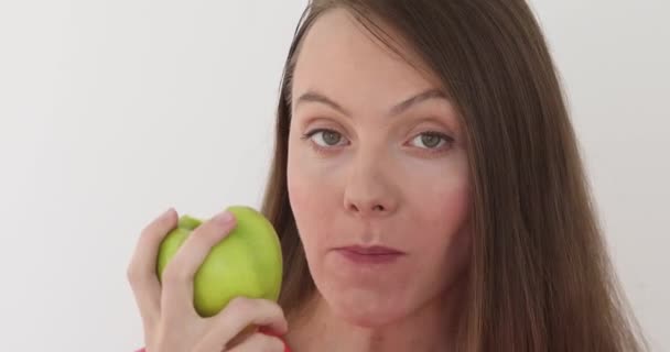 Young woman is eating a big green apple at white background. Healthy nutrition model eating fruit. Girl takes first bite and then offer bite to viewer and saying Wanna bite - Filmagem, Vídeo