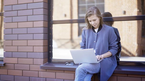 A young smiling girl is sitting on a sill typing in a laptop - 写真・画像