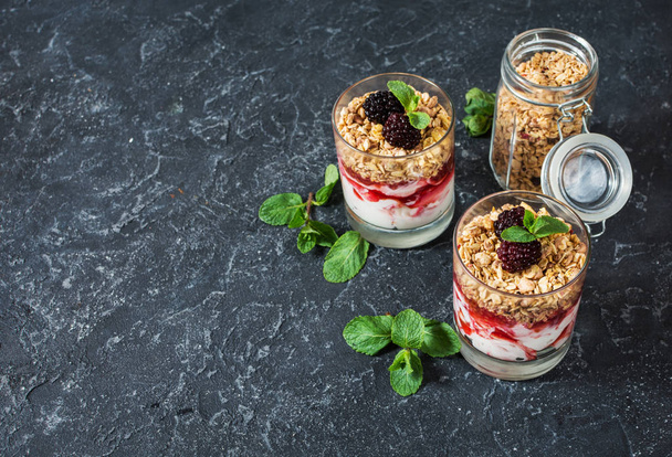 Healthy layered dessert with yogurt, granola, jam, blackberry in glass on stone background. Copy space - Zdjęcie, obraz