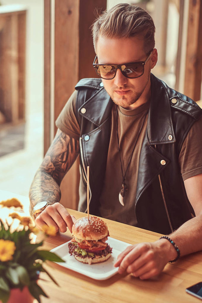 Knappe hipster met een stijlvol kapsel en baard zit aan een tafel in een café langs de weg, het drinken van een frisdrank op een warme dag. - Foto, afbeelding