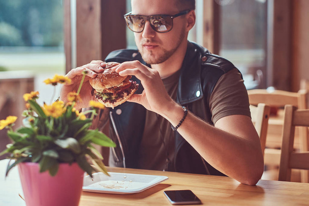 gutaussehender Hipster mit stylischem Haarschnitt und Bart sitzt an einem Tisch, entschließt sich, in einem Straßencafé zu essen und einen Hamburger zu essen. - Foto, Bild