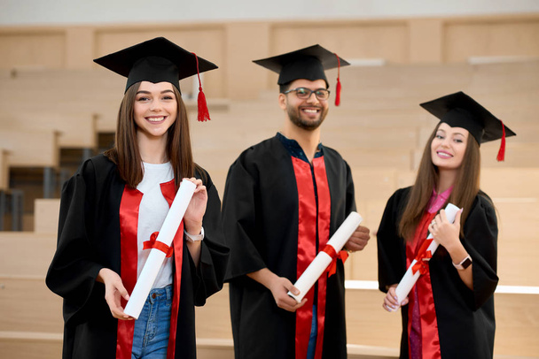Smiling graduates keeping diplomas in front of wooden desks. - Foto, Imagem