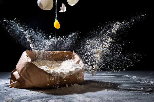 Bag with flour and egg flying in flour on black background. Flour splash. Cooking, baking concept - Photo, image