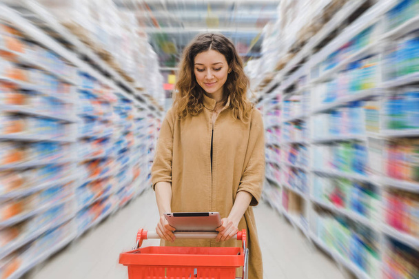 junge Geschäftsfrau mit Einkaufswagen in einem Supermarkt mit Blick auf ein Tablet zwischen den Regalen - Foto, Bild