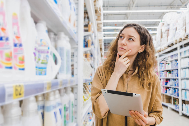 ビジネス女性がスーパーでタブレットを選択します家庭用化学薬品と買い物について考えています。 - 写真・画像