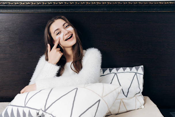 ospalý děvče v bílým pyžamu leží v posteli, brzy ráno, pod očima bílé skvrny, s úsměvem - Fotografie, Obrázek