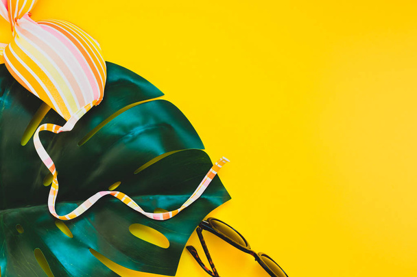Tropische Dschungelpalmen-Monsterblätter, Sonnenbrille und Badeanzug-Oberteil einer Frau auf leuchtend gelbem Papierhintergrund. Sommerferien kreatives flaches Lagekonzept Vorlage für Text - Foto, Bild