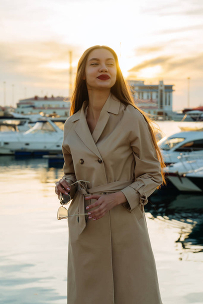 beautiful attractive girl with long hair and in a trendy beige coat is standing in the seaport and smiling - Photo, Image