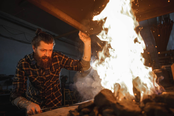Retrato de una profesión. Brutal herrero con barba enciende un fuego en el horno. Retrato a través de la llama
. - Foto, imagen