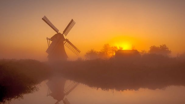 Charakteristický historickým holandský větrný mlýn podél koryta v mokřadu poldru na mlhavé ráno září v Nizozemsku - Fotografie, Obrázek