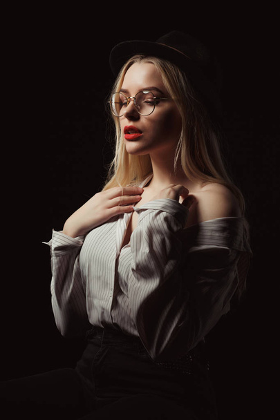 Romantický blondýnka ve brýle a klobouk, nosí košili s nahá ramena, pózování s dramatické světlo v ateliéru - Fotografie, Obrázek
