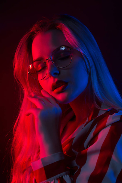 Closeup portrait of pretty young woman in glasses wearing striped blouse, posing with red and blue light on her face - Foto, Imagen