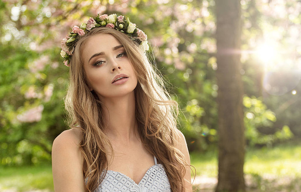 Beautiful caucasian blonde woman with long hair and flowers on head relaxing in blooming garden. Summer time. Leisure. Girl wearing fashionable dress. - Zdjęcie, obraz