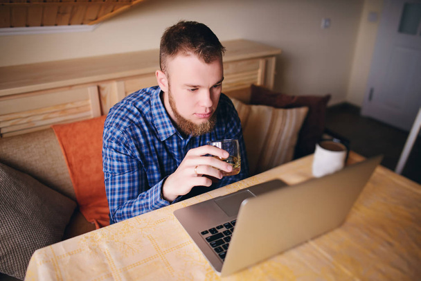 junger Profi surft auf seinem Laptop im Internet und trinkt Whiskey aus einem Glas. - Foto, Bild