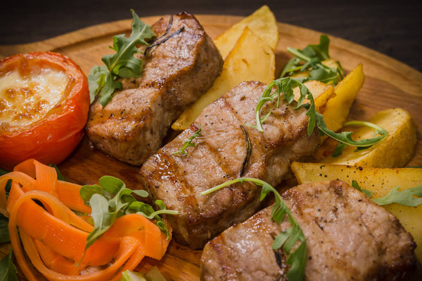 Три свиных стейка с жареным картофелем и овощами на деревянном фоне
 - Фото, изображение