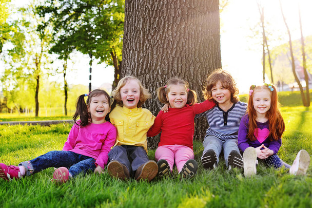 a group of small children in colorful clothes embracing sitting on the grass under a tree in a park laughing and smiling. - Фото, изображение