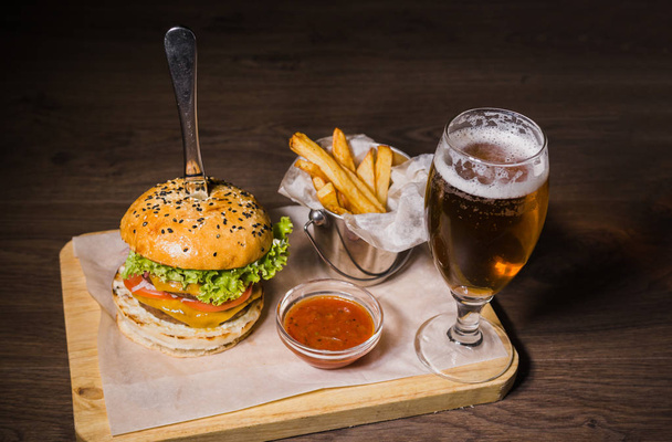 Burger with fried potatoes and tomato sauce on a wooden stand with a glass of beer - Zdjęcie, obraz