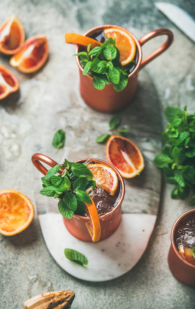 Blood orange Moscow mule alcohol cocktails with fresh mint leaves and ice in copper mugs on board in center over grey concrete background - Valokuva, kuva