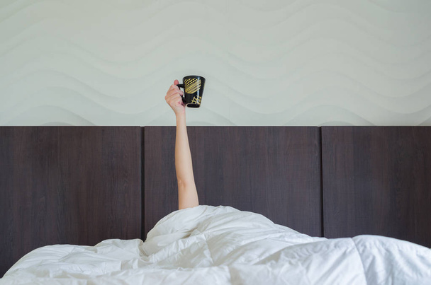 Drinking coffee early tomorrow. Woman in bed, under duvet, with arm raised up holding coffee cup. - Photo, image