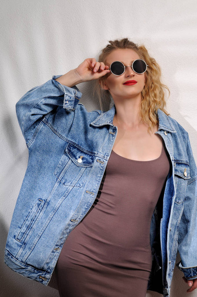 Fashion portrait of beautiful woman wear sunglasses in dress and jeans jacket on white wall  - 写真・画像