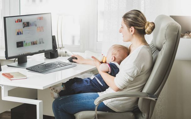 Молода жінка працює вдома з комп'ютером і годує свою дитину молоком
 - Фото, зображення