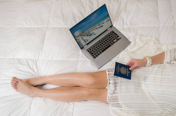Great concept of vacation planning, woman planning the holidays, passport in hand, computer, legs crossed, seen from above, in bed. - Foto, afbeelding
