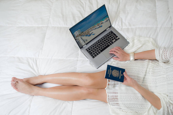 Geweldig concept van vakantie planning, vrouw planning van de vakantie, paspoort in de hand, computerwetenschappen, benen gekruist, van bovenaf gezien in bed. - Foto, afbeelding