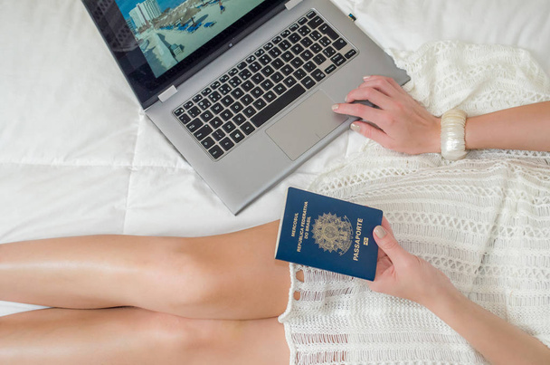 Great concept of vacation planning, woman planning the holidays, passport in hand, computer, legs crossed, seen from above, in bed. - Foto, afbeelding