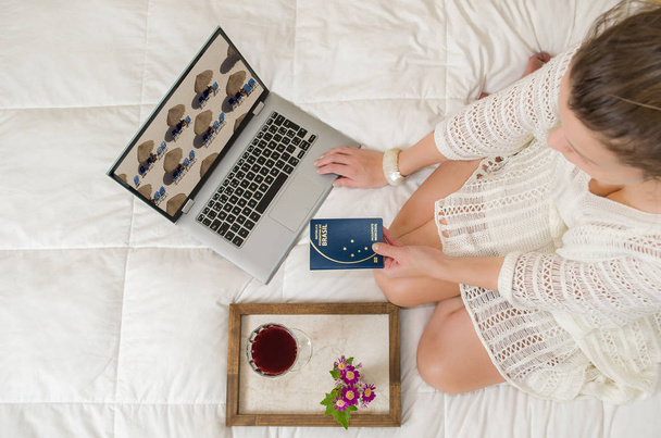 Great concept of vacation planning, woman planning the holidays, passport in hand, computer, legs crossed, seen from above, in bed. - Photo, image