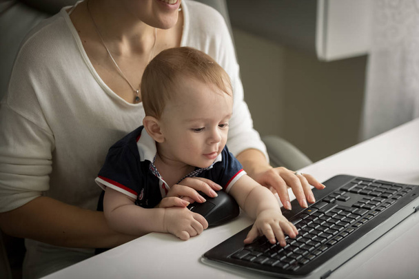 Portrait of 1 year old baby boy sitting with mother behind office desk and typing on computer keyboarrd - Photo, Image