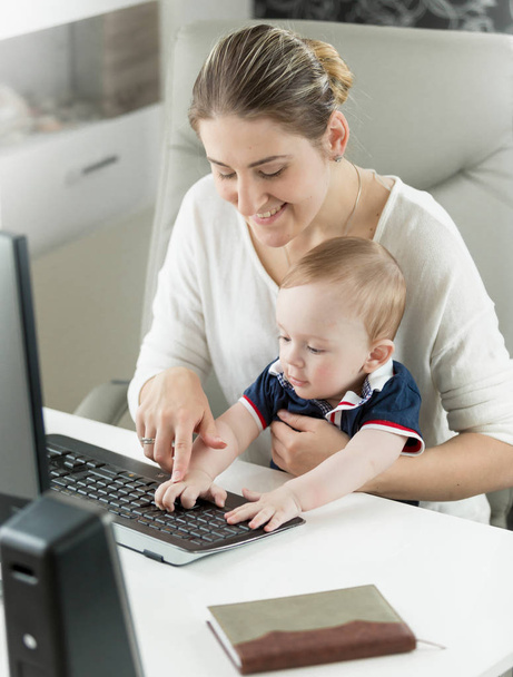 Портрет улыбающейся молодой женщины, обучающей своего маленького сына с помощью компьютера в офисе
 - Фото, изображение