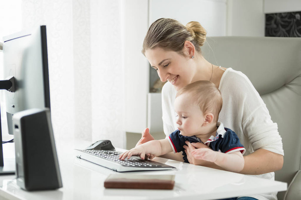 コンピューターを使用して、キーボードで入力する彼女の赤ちゃんの息子を教える若い母の肖像画 - 写真・画像