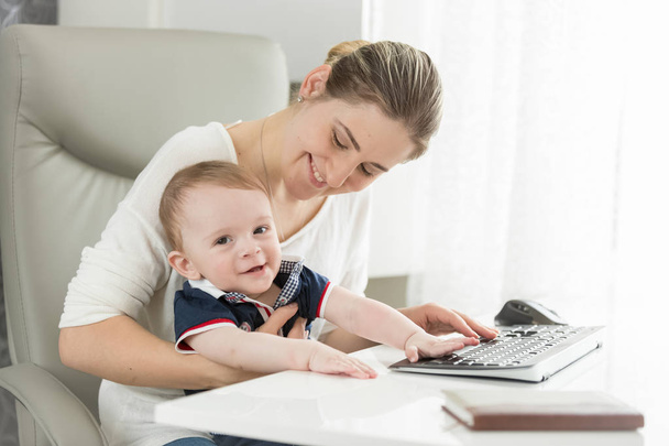 Feliz madre joven con hijo bebé sentado detrás del escritorio en la oficina en casa
 - Foto, imagen