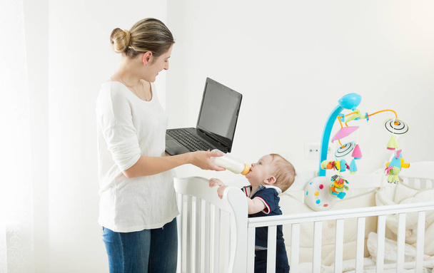 Portrait of young busy woman working on laptop and giving milk to her baby son - Фото, изображение