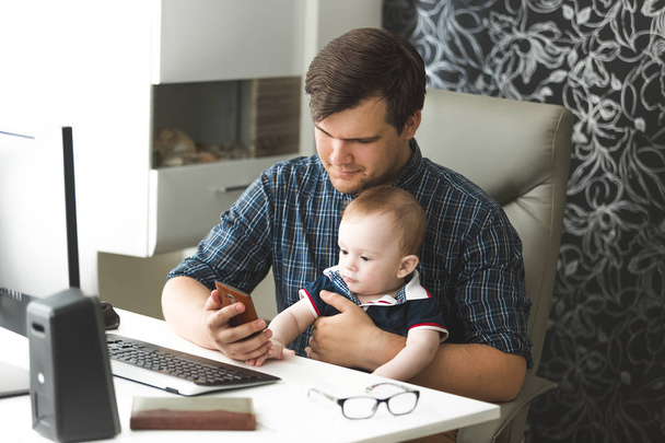 Getinte portret van jonge zelfstandigen man thuis werken met de computer en mobiele telefoon en op zoek naar zijn zoon baby - Foto, afbeelding