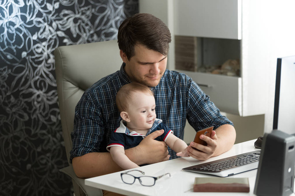 オフィスで彼の赤ん坊の息子と座っていると、スマート フォンを見て流行に敏感な若い男性の肖像画 - 写真・画像