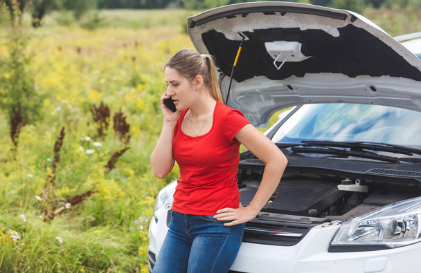 Portrait of young upset woman leaning on broken car in field and calling for help - Foto, Imagen