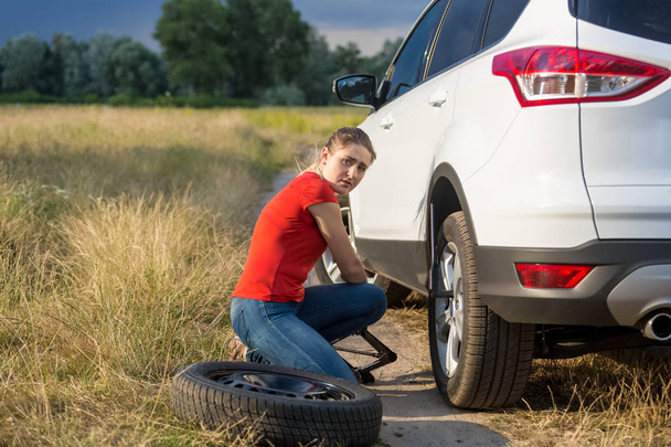 Giovane donna sconvolta sollevare la sua auto rotta con jack per cambiare pneumatico a terra su strada di campagna
 - Foto, immagini