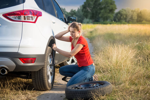 Verärgerte junge Frau schaut auf den platten Reifen ihres Autos auf Landstraße - Foto, Bild