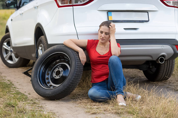 Retrato de mulher jovem apoiada no carro com pneu furado no campo
 - Foto, Imagem