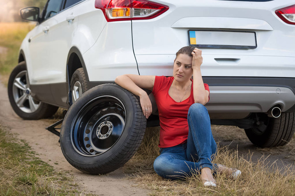 Nuori nainen istuu auton vieressä puhjennut rengas pellolla ja odottaa apua
 - Valokuva, kuva